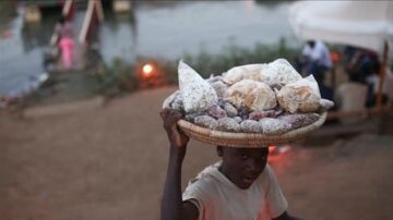 طفل يحمل سلة كبيرة مليئة بالخبز والحلويات على رأسه، يقف بالقرب من نهر. في الخلفية، يمكن رؤية أشخاص آخرين وبعض الأضواء. تعكس الصورة الحياة اليومية والتجارة المحلية في المنطقة.