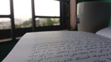 كتاب مفتوح على طاولة بجانب نافذة، يظهر نص باللغة العربية، مع كوب مشروب في الخلفية، وأفق المدينة واضح من النافذة.