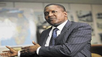 رجل أعمال يتحدث بجدية في مقابلة، يرتدي بدلة رمادية مخططة، مع خلفية تحتوي على خريطة أو صور. يعبّر عن أفكاره بيديه.