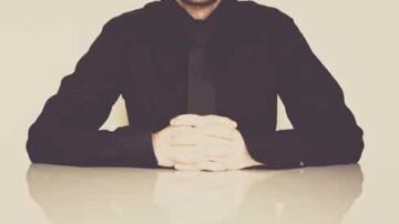 رجل يرتدي قميصًا أسود ويجلس على طاولة، يضع يديه معًا أمامه. الخلفية بسيطة بلون فاتح، مما يبرز مظهره الجاد.