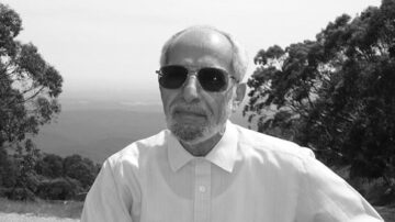 رجل مسن يرتدي نظارات شمسية وقميص أبيض، يجلس في منطقة طبيعية خضراء مع جبال في الخلفية. الصورة بالأبيض والأسود تعكس أجواء هادئة.