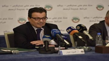 رجل يجلس على طاولة مؤتمرات يتحدث أمام ميكروفون، محاطاً بأجهزة إعلامية متعددة، مع شعار جهة رسمية خلفه.