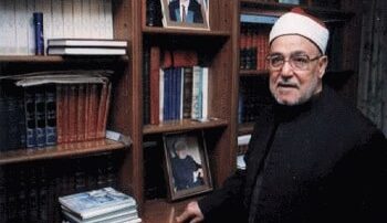 رجل مسن يرتدي عمامة بيضاء يقف بجانب مكتبة مليئة بالكتب، مع صور مؤطرة على الرفوف. يظهر في الخلفية مجموعة من الكتب الإسلامية والكتب الأخرى.