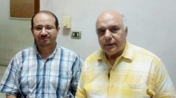 رجلان يجلسان معًا في غرفة داخلية، أحدهما يرتدي قميصًا مخططًا والآخر قميصًا صفراء. يبدو كلاهما في منتصف العمر، مع تعابير ودودة. الجدران خلفهم باللون الأبيض ويظهر جهاز تحكم في الحائط.