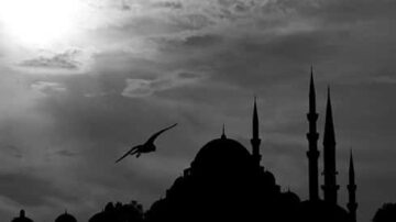 صورة بالأبيض والأسود تظهر silhouette لمسجد مع قباب ومآذن، بينما تحلق طائر في السماء المظلمة. السماء تحتوي على سحب مع شمس خافتة في الأفق، مما يضيف جوًا دراماتيكيًا.