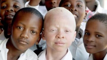 مجموعة من الأطفال يبتسمون، مع التركيز على طفل ذو بشرة فاتحة وشعر أشقر في المقدمة، بينما يقف بجانبه أطفال آخرون بملامح متنوعة. جميعهم يرتدون قمصانًا بيضاء، ويظهرون في بيئة مدرسية، مما يعكس التنوع والأمل في التعليم.