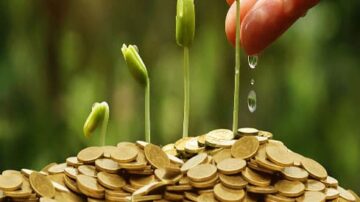 صورة تظهر يدًا تسقي براعم خضراء تنمو من كومة من العملات المعدنية. تعبر الصورة عن فكرة الاستثمار والنمو المالي.