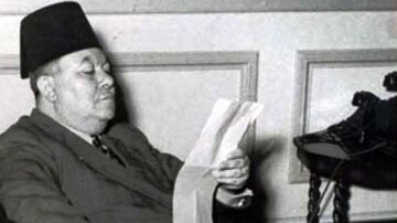 رجل يجلس في غرفة داخلية، يرتدي بدلة وقبعة تقليدية، ويقرأ ورقة. يظهر في الخلفية هاتف على طاولة. الصورة بالأبيض والأسود تعكس أجواء الزمن القديم.