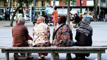 مجموعة من النساء بملابس تقليدية يجلسن على مقعد في ساحة عامة، يواجهن مشهد المدينة. تظهر في الخلفية أشخاص آخرون يتجولون، مع مبانٍ ومتاجر. الأجواء تعكس الحياة اليومية في المدينة.