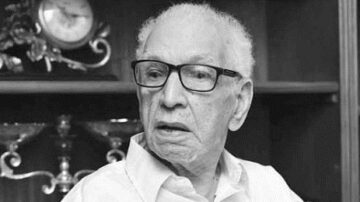 رجل مسن يرتدي نظارات، يجلس في غرفة مع أرفف وخلفية تحتوي على أشياء زينة وساعة. تعبر تعابيره عن الحكمة والخبرة.