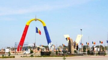 قوس ملون بألوان العلم الروماني في ساحة عامة، مع تمثال يحمل العلم ورموز ثقافية أخرى. السماء صافية والأرض مرصوفة، مما يبرز أهمية الموقع كمكان للاحتفالات والفعاليات.