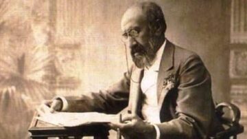 رجل مسن يجلس على طاولة، يرتدي بدلة رسمية، ويقرأ صحيفة. يبدو مركزًا وعميق التفكير، مع لحية وشعر رمادي. الخلفية غير واضحة، مما يبرز تفاصيل وجهه وتعابيره.