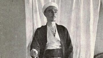 شاب يرتدي زي تقليدي مع عمامة ويقف أمام خلفية بيضاء. تعكس الصورة التراث الثقافي والفني.