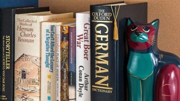 كتب مرصوصة على رف، تشمل "الأعمال الكاملة" و"قاموس أوكسفورد" و"الألمانية"، بالإضافة إلى تمثال قطة ملون يظهر على الجانب.