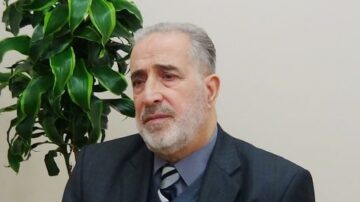 رجل مسن يرتدي بدلة رسمية ويجلس أمام نبات أخضر. يبدو عليه التفكير والتأمل.