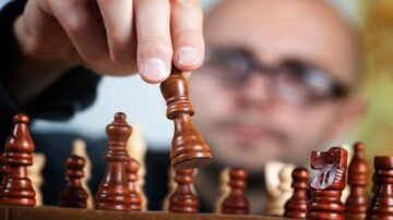 رجل يرتدي نظارات يرفع قطعة شطرنج خشبية من اللوحة، مع التركيز على حركته. تظهر قطع الشطرنج الأخرى في الخلفية، بعضها سليم وبعضها مكسور.