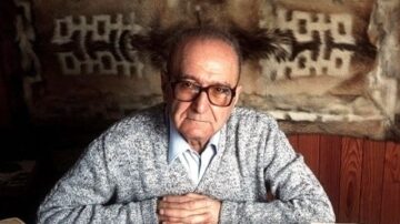 رجل مسن يرتدي نظارات وقميص أزرق تحت سترة رمادية مُزخرفة، يجلس أمام طاولة في غرفة ذات جدران خشبية مزينة بسجاد تقليدي. تعكس تعابير وجهه الحكمة والخبرة.