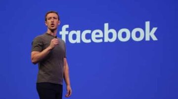 رجل يقف على خشبة المسرح يتحدث أمام شعار فيسبوك على خلفية زرقاء، يرتدي قميصاً رمادياً.