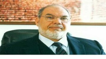 رجل مسن ذو لحية وشعر رمادي، يرتدي بدلة سوداء وقميص أبيض مع ربطة عنق. يجلس في مكتب، والخلفية تحتوي على جدار مغطى بالطوب. تعكس تعابير وجهه الثقة والجدية.