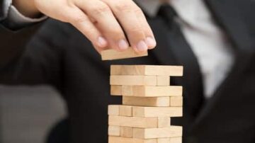 يدًا ترتفع لتضع قطعة من لعبة الجينجا فوق برج خشبي مرتفع، يرتدي الشخص بدلة رسمية. تركز الصورة على التوازن والتحدي في اللعبة.