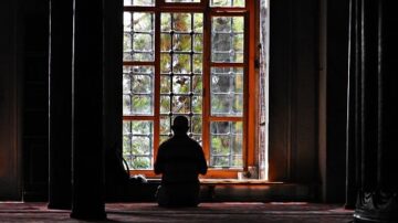 رجل جالس في الصلاة أمام نافذة كبيرة، حيث تتسلل أشعة الشمس من خلال الزجاج الملون، مما يخلق جوًا من الهدوء والسكينة في المكان.