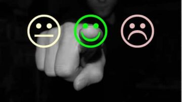 يد تشير بإصبعها إلى ثلاثة وجوه تعبيرية: وجه محايد باللون الأصفر، وجه مبتسم باللون الأخضر، ووجه حزين باللون الوردي. تعبر الصورة عن مشاعر مختلفة وتفاعل الإنسان مع العواطف.