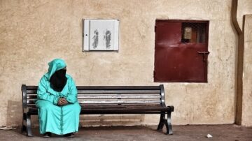 امرأة ترتدي جلباباً أخضر وتغطي وجهها، جالسة بمفردها على مقعد خشبي في مكان ذو جدران بيج. خلفها باب أحمر ولوحة بيضاء فارغة، تعكس أجواء هادئة وتفكير عميق.