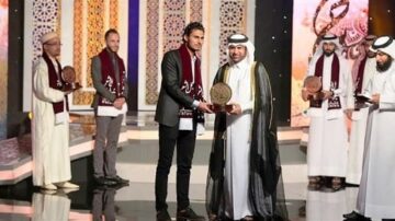 مراسم توزيع الجوائز في برنامج تلفزيوني، يظهر فيها فائز يتلقى جائزة من شخصية بارزة. في الخلفية، مجموعة من المكرمين يرتدون أزياء تقليدية. الإضاءة الساطعة والزينة العربية تبرز الأجواء الاحتفالية.