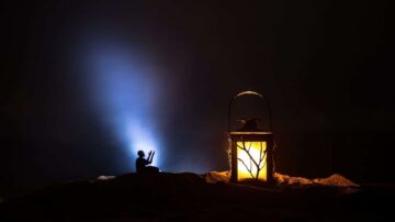 صورة تمثل شخصية صغيرة تجلس بجانب مصباح مضيء في بيئة مظلمة. يضيء المصباح بلون دافئ بينما تنبعث أشعة ضوء زرقاء في الخلفية، مما يخلق تباينًا جذابًا. تعكس الصورة أجواء من التأمل والسكينة.