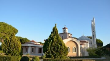 كنيسة محاطة بالأشجار الخضراء، تحت سماء زرقاء صافية. تظهر المعمارية التقليدية مع قبة وجرس، مما يبرز جمال المكان.