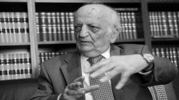 رجل مسن يتحدث بحماس، مرتديًا بدلة رسمية، جالس في مكتب محاط برفوف مليئة بالكتب القانونية. تعبير وجهه يعكس الجدية والاهتمام بالموضوع الذي يناقشه.