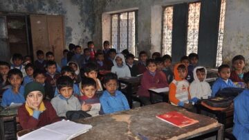 فصل دراسي مزدحم يحتوي على مجموعة من الأطفال يجلسون على المكاتب. الأطفال يرتدون ملابس متنوعة، وبعضهم يرتدي قبعات. الجدران متآكلة والنوافذ تحتوي على زجاج مكسور، مما يعطي انطباعًا عن بيئة تعليمية قديمة.