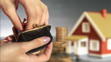 يد تظهر حقيبة نقود سوداء مفتوحة مع عملات معدنية في الخلفية، ومنزل صغير على شكل نموذج. الصورة تعبر عن مفهوم المال والادخار في مجال العقارات.