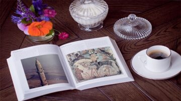 كتاب مفتوح على طاولة خشبية، يظهر صورة لمشهد فني. بجانبه فنجان قهوة أبيض وزهور ملونة، مع وعاء زجاجي يحتوي على كريمة.