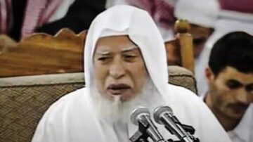 رجل مسن يرتدي عمامة بيضاء ولديه لحية طويلة، يتحدث أمام ميكروفونات في حلقة نقاش. خلفه يظهر عدد من الأشخاص في الخلفية.