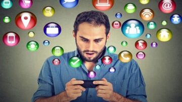 رجل شاب ينظر بدهشة إلى هاتفه الذكي، محاط بفقاعات ملونة تمثل رموز وسائل التواصل الاجتماعي والتطبيقات، مثل أيقونات الموقع، والمكالمات، والشبكات الاجتماعية، مما يعكس تأثير التكنولوجيا والتواصل الرقمي.