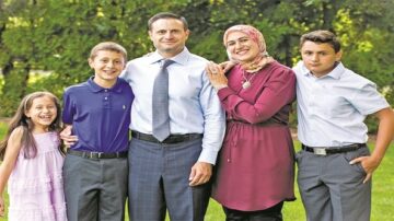 عائلة مكونة من خمسة أفراد تتوسطهم والدتهم، يقفون بابتسامات في حديقة. الأطفال ثلاثة، ولدين وبنت، يرتدون ملابس أنيقة. الأم ترتدي حجاباً وملابس طويلة، بينما الأب يظهر بشكل رسمي. الشجر والخضرة في الخلفية تضيف لمسة طبيعية للصورة.