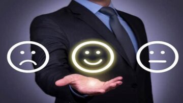 رجل يرتدي بدلة رسمية يحمل بيده ثلاثة رموز تعبيرية، واحد مبتسم وآخر حزين وثالث محايد، مع خلفية داكنة، مما يعكس موضوع التعبير عن المشاعر في بيئة العمل.