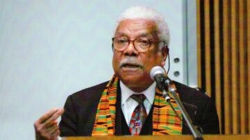 رجل مسن يتحدث في ميكروفون، يرتدي سترة سوداء مع وشاح كُنعاني ملون، يظهر خلفه خلفية بسيطة. يُظهر الصورة تعبيرات وجهه الجادة أثناء إلقاء حديث مهم.