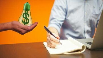 يد تمسك بمصباح كهربائي أخضر يحمل رمز الدولار، بينما شخص يجلس أمام كمبيوتر محمول ويكتب في دفتر ملاحظات. الخلفية برتقالية، مما يضيف إحساسًا بالطاقة والابتكار. هذه الصورة تمثل فكرة الإبداع في الأعمال والتفكير المالي.