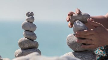 أيدي تكدس الأحجار الرمادية على شاطئ البحر مع وجود كومة من الأحجار في الخلفية. تعكس الصورة النشاط الإبداعي والتوازن في بناء الأشكال الحجرية.