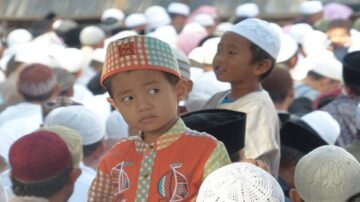 طفل يرتدي قبعة ملونة ونظرة فضولية وسط حشد من الناس في ملابس تقليدية، يعبر عن لحظة من الفعالية الثقافية أو الدينية.