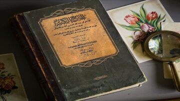 كتاب تاريخي قديم باللغة العربية مع غلاف جلدي، موضوع على طاولة بجانب عدسة مكبرة وصور زهور ملونة.