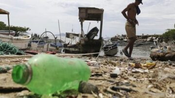رجل يسير على شاطئ ملوث بالنفايات، مع زجاجة بلاستيكية خضراء في المقدمة، وقوارب صيد في الخلفية، تعكس تأثير التلوث على البيئة البحرية.