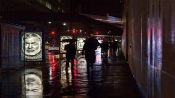 مشهد ليلي لشارع حضري مع أشخاص يحملون مظلات. تظهر لوحات إعلانات مضيئة على الجانبين، تعرض صوراً لأشخاص. تعكس الأضواء والمطر على الرصيف، مما يخلق أجواء درامية.