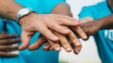 أيدي متعددة تتداخل فوق بعضها البعض، تعبيرًا عن التعاون والتضامن، جميع الأيدي ترتدي قمصانًا زرقاء، في خلفية غير واضحة تعكس نشاطًا جماعيًا.