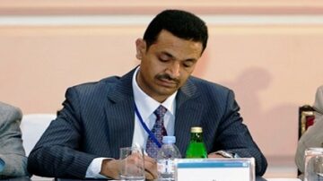 رجل يرتدي بدلة رسمية يكتب ملاحظات على ورقة خلال مؤتمر أو اجتماع، مع زجاجة مياه ووعاء زجاجي على الطاولة.