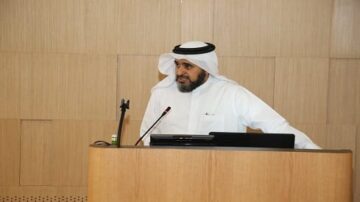 رجل يرتدي الزي التقليدي العربي يتحدث في مؤتمر، يقف خلف منصة خشبية مع ميكروفون وجهاز كمبيوتر محمول. تظهر خلفه جدران خشبية.