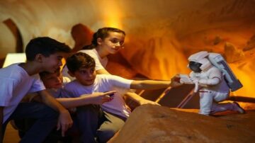 أربعة أطفال يرتدون قمصان بيضاء يتفاعلون مع تمثال لرائد فضاء في بيئة داخلية تشبه الكهف، مضاءة بألوان دافئة. الأطفال يظهرون اهتمامًا واستكشافًا للتمثال، مما يعكس روح الفضول والابتكار.