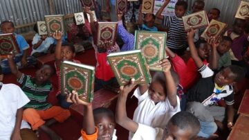 مجموعة من الأطفال في فصل دراسي, يرفعون كتب القرآن بفرح. تظهر الوجوه المتحمسة والحماس في الأجواء. الفصول الدراسية بسيطة، مع جدران معدنية. تعكس الصورة أهمية التعليم الديني في المجتمعات.